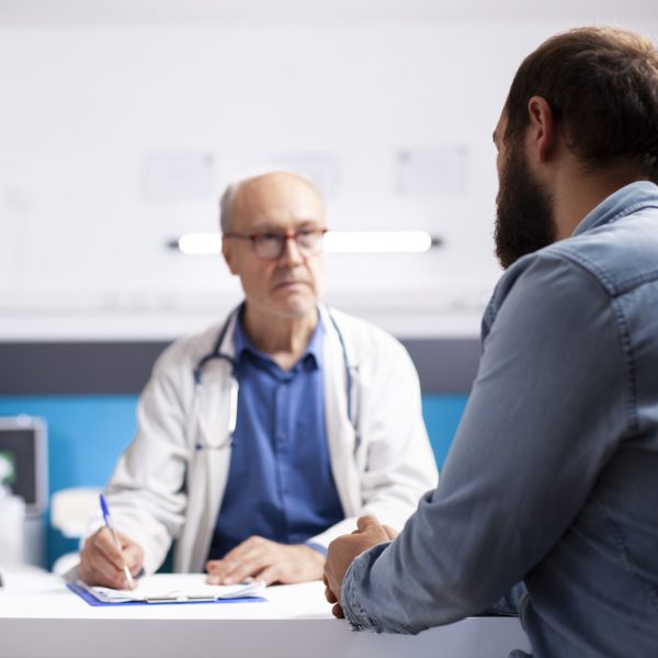 Elderly healthcare specialist in medical office talks with male patient, taking notes on his clipboard. Professional old physician listening to health concerns of young man during clinic appointment.